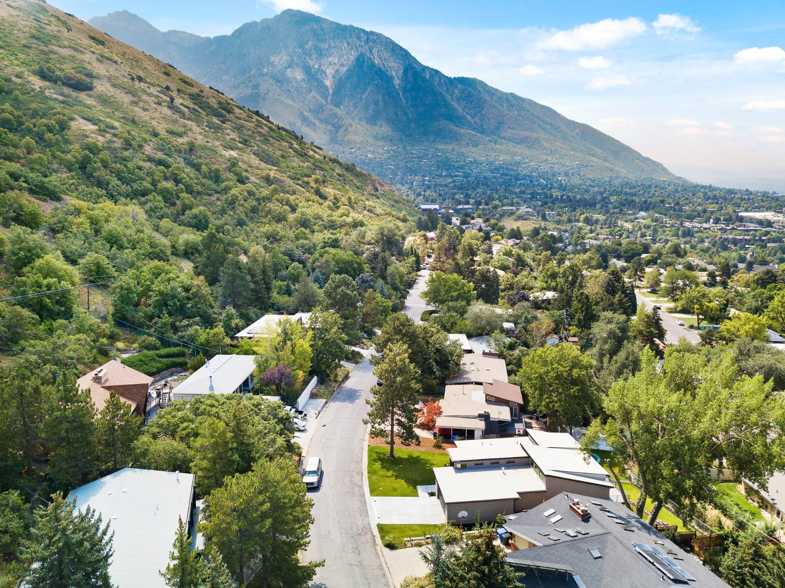 Elevated view of homes, lawns, and tree-lined streets at the foot of steep mountains under a blue sky with scattered clouds.